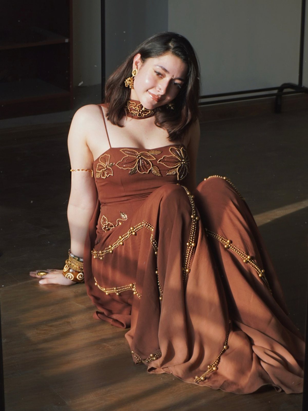 Woman in a brown embroidered gown with gold jewelry sitting on a wooden floor