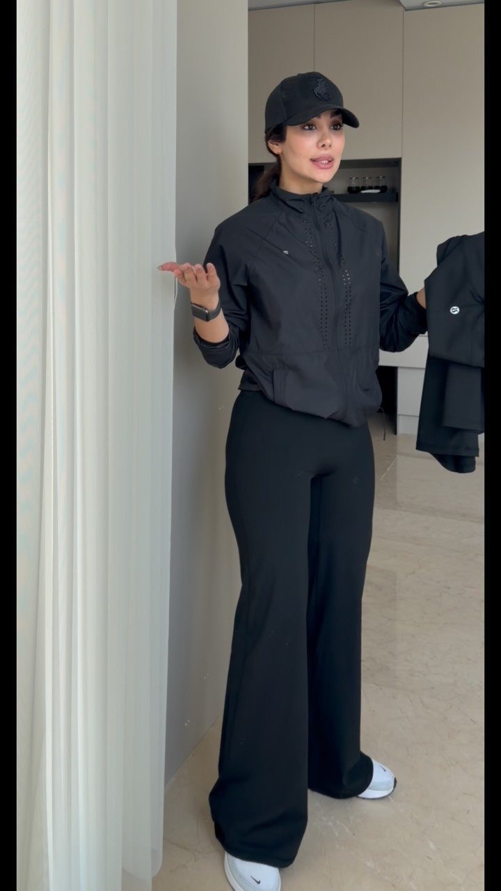 Woman wearing black baseball cap, black zip-up jacket, and black wide-leg athletic pants.