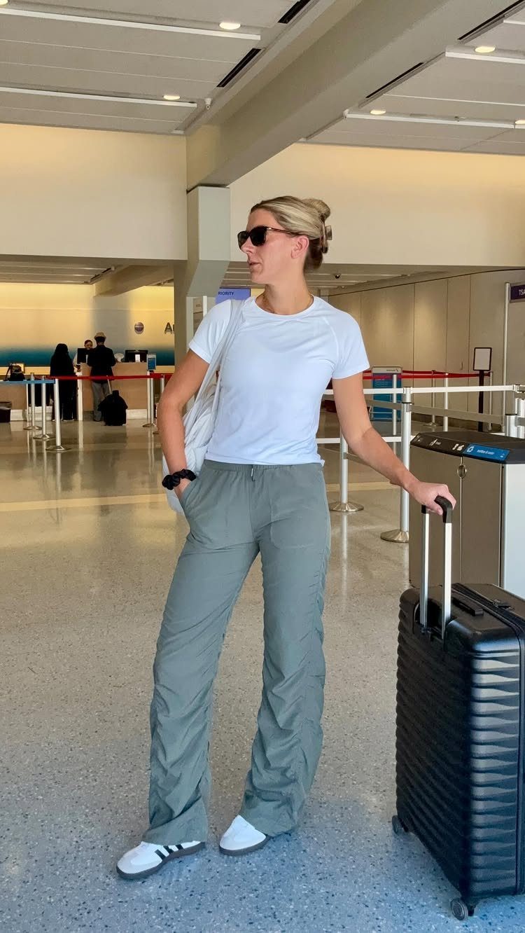 Woman wearing white t-shirt and olive green wide-leg pants standing with a suitcase.