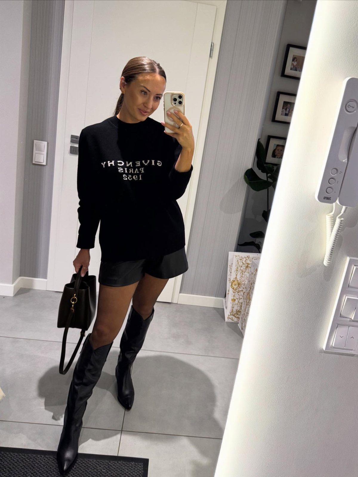 Woman in black sweatshirt and knee-high boots taking a mirror selfie indoors.