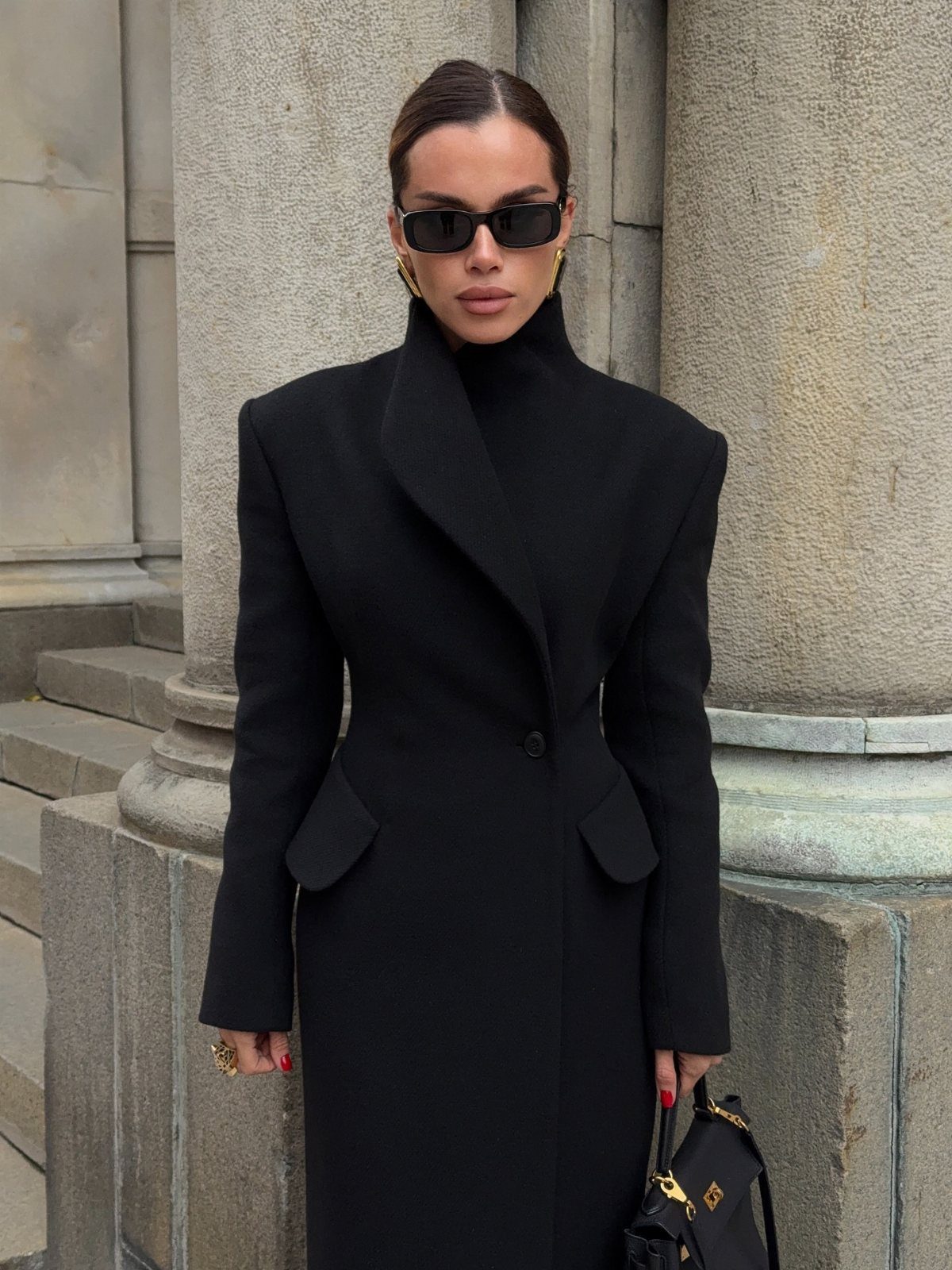 Woman in a long black structured coat and turtleneck standing by stone columns.