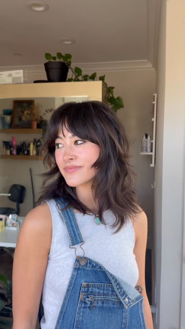 16 Shoulder Length Layered Hair Ideas for Every Texture 4 Woman with dark brown choppy layered hair and full fringe bangs wearing overalls.