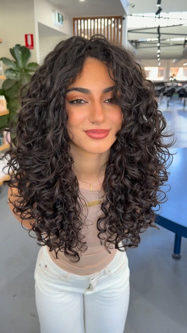 16 Shoulder Length Layered Hair Ideas for Every Texture 16 Woman with voluminous curly dark brown shoulder length layered hair in a salon.
